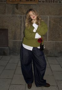 Jeune femme appuyée contre un mur de pierre, portant un pull vert, une chemise blanche, un pantalon large foncé, et tenant un petit sac avec un chouchou rouge.