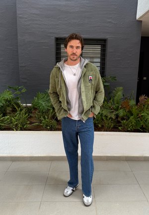 Chaqueta de pana verde oliva con capucha gris, camiseta blanca, jeans azules y zapatillas grises con acentos negros, todo en un fondo de follaje verde.