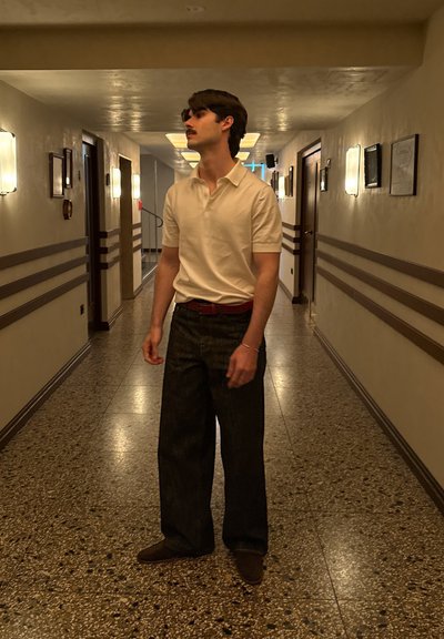 Camisa polo blanca, pantalones de mezclilla oscuros de campana, cinturón marrón y zapatos. De pie en un pasillo bien iluminado con luminarias decorativas.