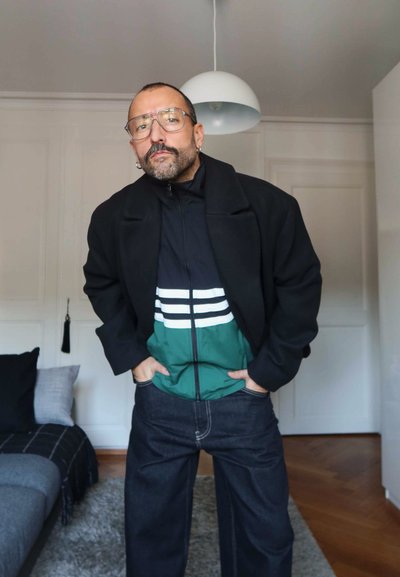 Hombre con gafas, barba y pendientes, vistiendo un abrigo negro sobre una chaqueta con cremallera verde y negra, de pie en el interior con las manos en los bolsillos.