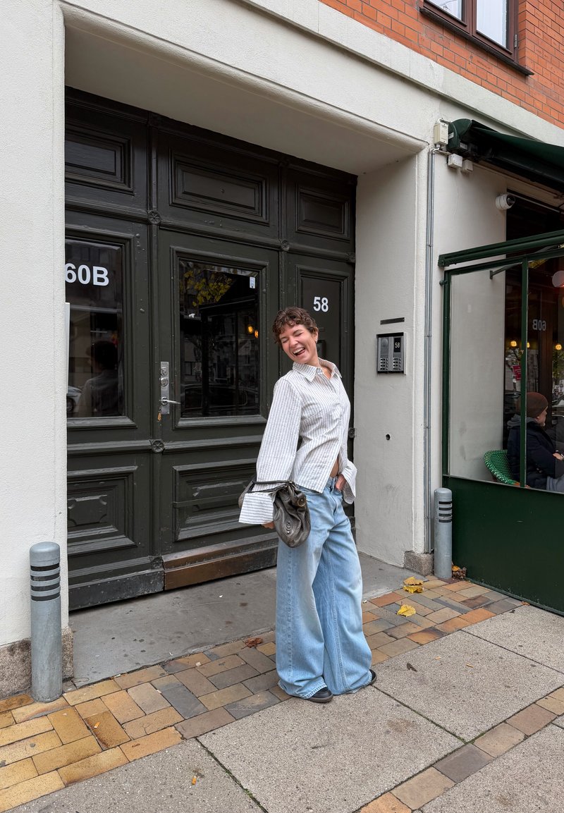 Jeans oversized de ganga azul claro e uma camisa branca às riscas com botões. A pessoa segura uma pequena mala cinzenta, estando de pé perto da entrada verde de um café.
