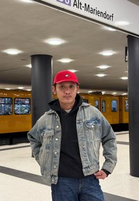 Veste en denim par-dessus un sweat à capuche noir, jean bleu et casquette rouge avec un logo. Station de métro avec un train orange en arrière-plan.