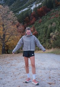 Persona sporta apģērbā pielāgo jaku uz lapām pārklāta ceļa, ko ieskauj rudens koki un zaļi pakalni.