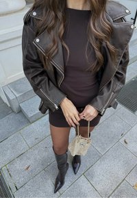 Žena u tamnosmeđem kožnom sakou i haljini, drži malu bež tkanu torbicu, stoji na sivom kamenitom pločniku.