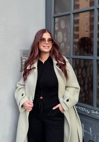 Krēmkrāsas pārspīlēts mētelis, melna ribota blūze, melni bikses un apaļi saules brilles; gari viļņaini sarkani mati; stāv pie stikla loga un telekomunikāciju ierīces.