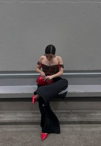 Moteris sėdi ant suolo, vilkėdama liūto rašto off-the-shoulder palaidinę, juodas sportines kelnes su baltomis juostelėmis ir raudonus smailius aukštakulnius. Rankose laiko raudoną rankinę.