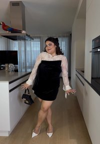 Vestido mini de veludo preto com mangas longas de renda, sapatos brancos de salto pontudo e uma pequena bolsa preta. Fundo de cozinha moderna com chão de madeira.
