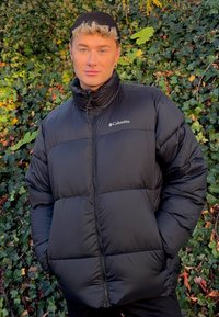 Svart pufferjacka med hög krage, en dragkedja fram och Columbia-logotyp på vänster bröst. Texturerad tyg och vadderat mönster.