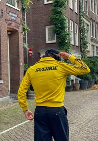 Κίτρινο μπουφάν γυμναστικής με την ένδειξη "SVERIGE" τυπωμένη σε ναυτικό χρώμα στην πλάτη, με ριγέ μπλε λεπτομέρειες στα μανίκια. Ορατό μαύρο παντελόνι.