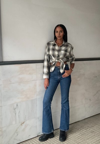 Camisa de cuadros con un nudo en el frente, combinada con jeans acampanados de talle alto en azul medio y botas de tobillo negras. Texturas suaves y modernas.