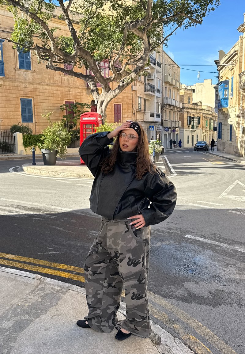 Frau in Tarnhosen und schwarzer Jacke steht an einer Straßenecke mit der Hand am Kopf, roter Telefonzelle und Baum im Hintergrund, sonnige städtische Umgebung.