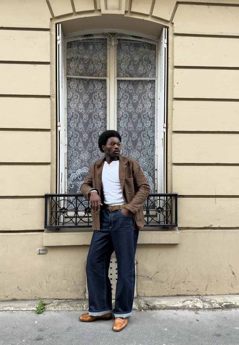 Man in een bruin jasje, wit hemd, donkere jeans en lichtbruine schoenen leunend tegen een beige gebouw met een hoog raam bedekt met kanten gordijnen.