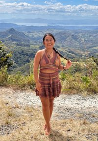 Femme souriante pieds nus portant un ensemble deux pièces marron à motifs, tenant ses cheveux tressés, avec un paysage montagneux et un ciel bleu en arrière-plan.