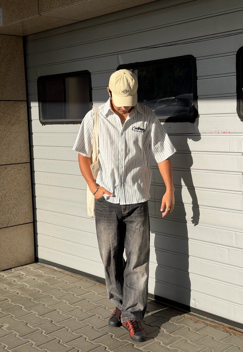 Homme portant une casquette beige, une chemise à manches courtes rayée, un jean noir et des chaussures rouges marchant devant une porte de garage au soleil.