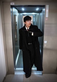 Homme portant des lunettes et un long manteau noir sort d'un ascenseur moderne, ajustant son manteau avec une main gantée.