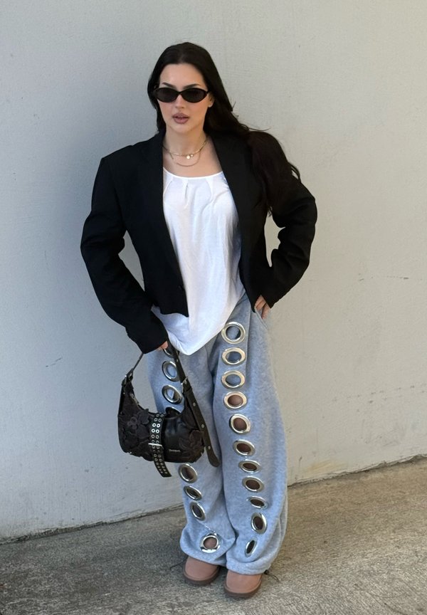 Donna che indossa blazer nero, top bianco, pantaloni grigi con grandi anelli metallici, occhiali da sole, tiene una borsa nera, in piedi contro una parete liscia.