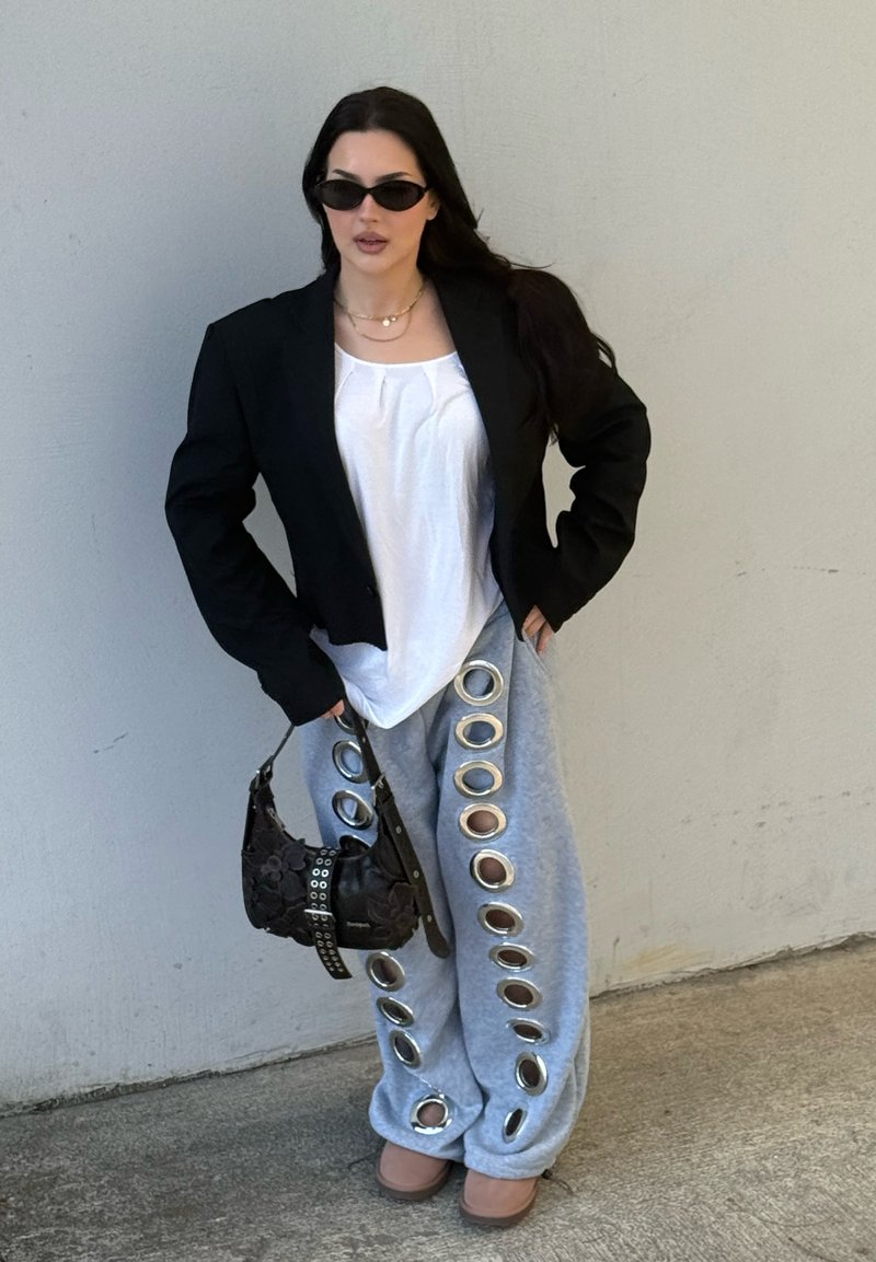 Femme portant un blazer noir, un top blanc, un pantalon gris avec de grands anneaux métalliques, des lunettes de soleil, tenant un sac à main noir, debout contre un mur uni.