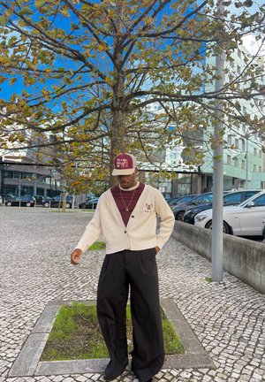 Hombre con cárdigan beige, camisa burdeos y gorra, de pie cerca de un árbol cubierto de hojas en una acera de adoquines en una zona de aparcamiento urbana.