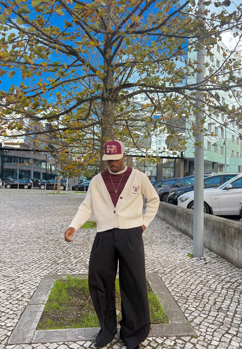 Man i beige kofta, vinröd skjorta och keps som står vid ett lövbeklätt träd på en kullerstensgång i ett stadsområde med parkering.