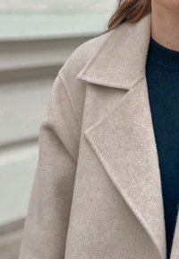 Világos bézs gyapjúkabát, kivágott gallérral, textúrált anyagból, laza szabású. Sötétkék pulóver fölött viselve.