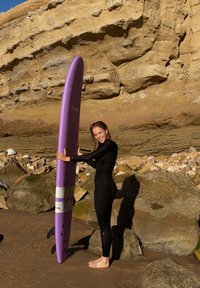 Femme en combinaison noire debout pieds nus sur une plage de sable tenant une grande planche de surf violette près d'une falaise rocheuse sous le soleil.