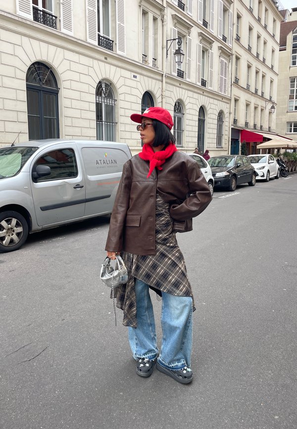 Bruine leren jas, rode sjaal, geruite asymmetrische jurk, brede blauwe jeans, zwarte platform schoenen, zilveren tas en donkere zonnebril.