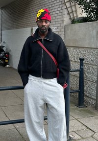 Maglione nero a zip, pantaloni della tuta grigi e un berretto a fantasia multicolore. Borsa a tracolla rossa e sfondo in mattoni discreti.
