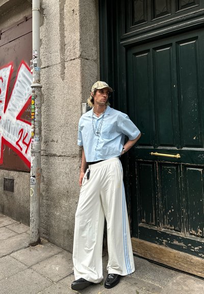 Camisa de botones de manga corta azul claro, pantalones anchos blancos con rayas azules, zapatos negros y una gorra de camuflaje, complementada con cuentas.