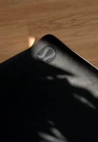 Zwarte yogamat met afgeronde hoeken, met een subtiele reliëf logo. Gladde, matte textuur, gedeeltelijk verlicht door zonlicht.
