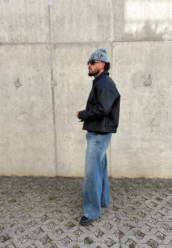 Hombre con chaqueta negra, vaqueros azules, gorro azul con símbolos y gafas de sol, de pie de lado contra una pared de hormigón sobre un pavimento con patrón.