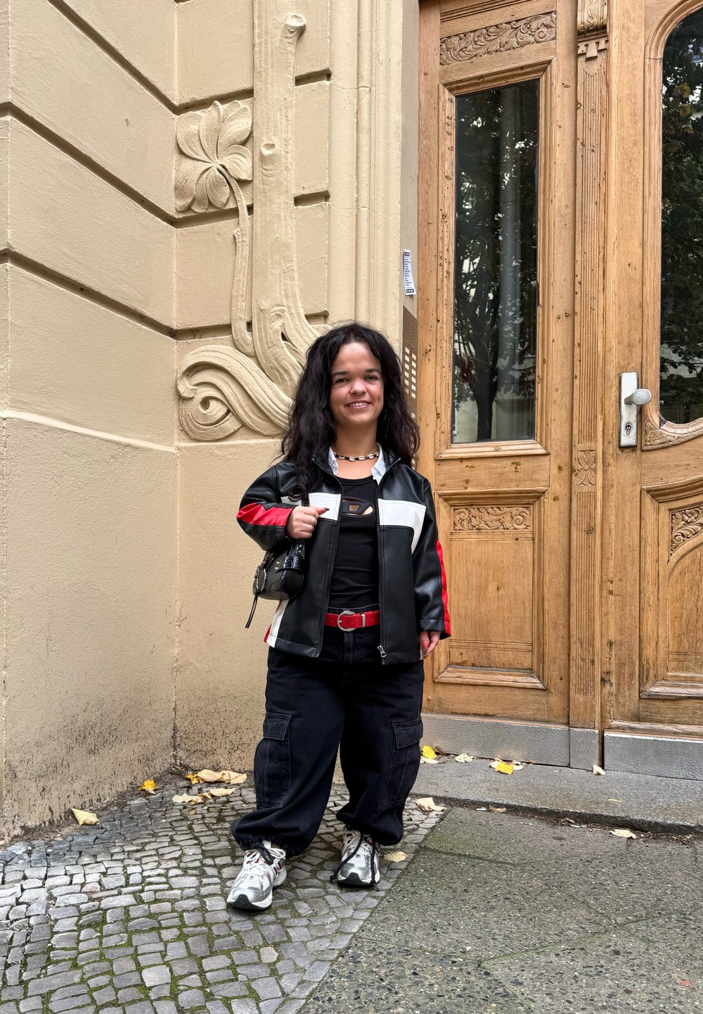 Giacca in pelle nera e rossa con accenti bianchi, maglietta nera, cintura rossa, pantaloni neri larghi, sneakers grigie e bianche, in piedi vicino a una porta di legno.