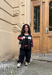 Giacca in pelle nera e rossa con accenti bianchi, maglietta nera, cintura rossa, pantaloni neri larghi, sneakers grigie e bianche, in piedi vicino a una porta di legno.