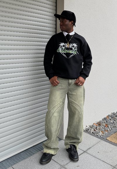 Sudadera negra con diseño gráfico, pantalones anchos verde claro y zapatos negros con borlas. Hombre de pie contra una pared blanca texturizada.