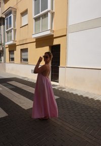 Μακρύ ροζ φόρεμα χωρίς μανίκια με ανοιχτή πλάτη, φορεμένο σε λιθόστρωτο δρόμο. Ο/Η φορέας κρατά γυαλιά ηλίου, με τον ήλιο να ρίχνει σκιές.