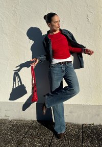 Haut rouge à manches longues sous une veste en jean, jean bleu évasé et chaussures pointues marron. Tenant un sac à main rouge. Ombre projetée sur un mur.