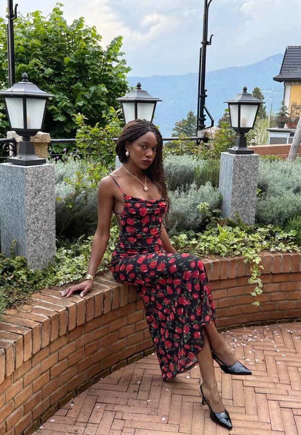 Kvinde i en sort kjole med røde roser, der sidder på en buet mur af mursten omgivet af lamper, grønt og udsigt til fjerne bjerge.