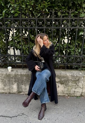 Mujer con cabello largo y rubio sentada con las piernas cruzadas en un banco de piedra junto a una verja de hierro forjado negra, vistiendo un abrigo negro y botas marrones.