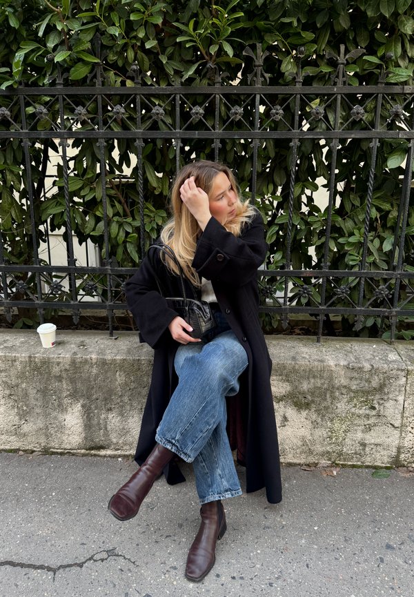 Femme aux longs cheveux blonds assise en tailleur sur un banc en pierre près d'une clôture en fer forgé noire, portant un manteau noir et des bottes marron.