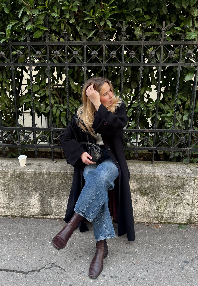 Žena s dlouhými blond vlasy sedí na kamenné lavičce přeskládaná, vedle černého kovaného železného plotu, oblečená v černém kabátě a hnědých botách.