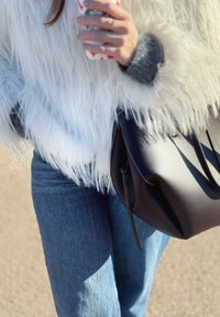 Personne portant une veste en fausse fourrure blanche et un jean bleu, tenant une tasse à motif festif et portant un sac en cuir noir.