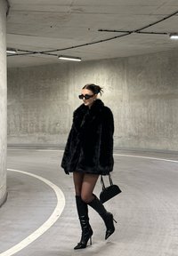 Crni kaput od umjetnog krzna sa širokim ovratnikom, uparen s crnim stiletto čizmama iznad koljena, crnom torbicom i tamnim naočalama; glatke teksture.