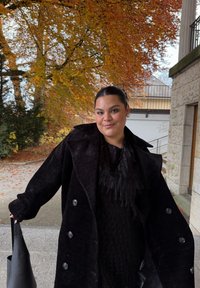 Černý manšestrový kabát s velkými knoflíky, nošený přes černý top s třásněmi. Drží černou tašku tote; na pozadí jsou podzimní listy.