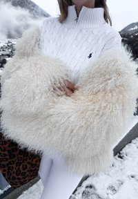 Personne portant un pull en maille torsadée blanc et une grande écharpe crème duveteuse, debout en plein air dans un décor de montagne enneigée.