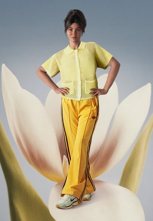 Camisa de botones amarilla de malla con bolsillos, combinada con pantalones deportivos amarillos con rayas negras. Zapatillas de deporte en verde claro con acentos grises.