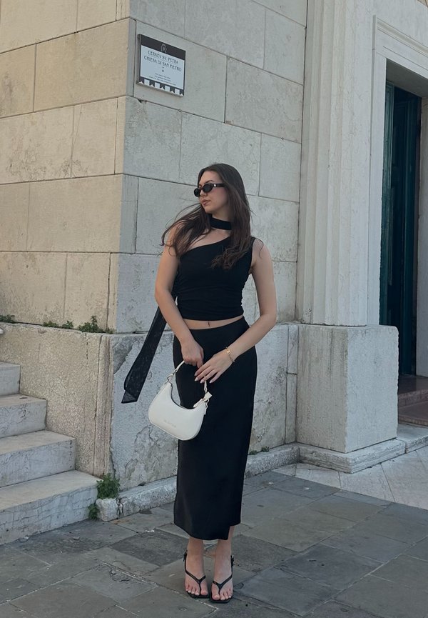 Schwarzes, tailliertes Crop-Top mit einem Rüschendetail, kombiniert mit einem schwarzen Midi-Rock. Trägt schwarze Sonnenbrillen und hält eine weiße Handtasche.