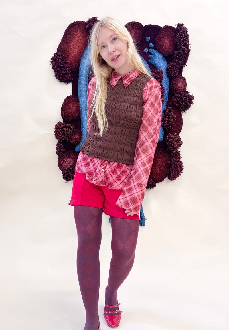 Femme blonde portant un gilet marron, une chemise à carreaux rose, un short rouge, des collants à motifs et des chaussures rouges, se tenant devant une œuvre murale texturée.