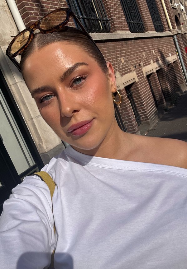 Giovane donna con capelli tirati indietro indossa orecchini a cerchio dorati e occhiali da sole sulla testa, vestita con un top bianco scollato sulle spalle all'aperto.