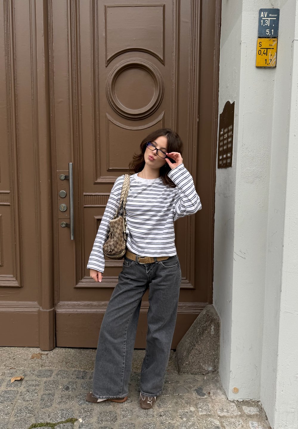 Chemise à manches longues rayée blanche et marine, associée à un jean denim gris ample. Sac à main marron et porte ronde en bois en arrière-plan.