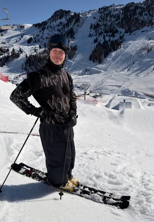 Hombre con conjunto de esquí negro, casco y gafas, de pie sobre esquís en una pendiente nevada con una estación de esquí montañosa al fondo.