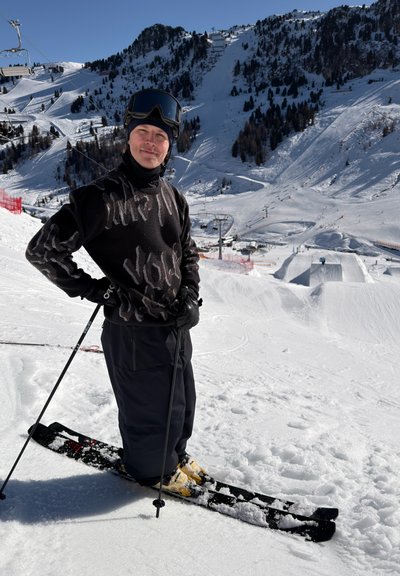 Hombre con conjunto de esquí negro, casco y gafas, de pie sobre esquís en una pendiente nevada con una estación de esquí montañosa al fondo.